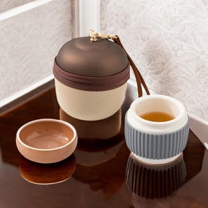 May include: A brown and beige travel tea set with a gray and white insulated teacup. The teacup has a lid and a silicone sleeve. The set is on a brown wooden surface.