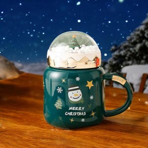 May include: Green ceramic mug with a snowman and Christmas tree design. The mug has a clear dome lid with a small Christmas tree inside. The mug has a handle with the word "LOVE" on it. The mug says "Merry Christmas".