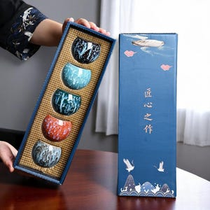 May include: A set of five small ceramic teacups in a blue box with a gold interior. The cups are decorated with different blue, green, and orange patterns. The box has the Chinese characters "匠之作" printed on the front.