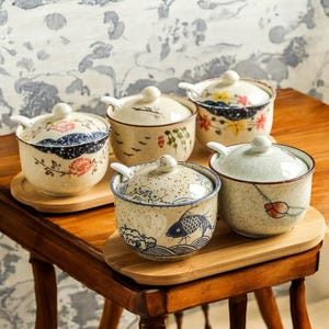 Puede incluir: Conjunto de cinco tarros de condimentos de cerámica con tapas y cucharas. Los tarros tienen diferentes diseños florales y geométricos en tonos azules, verdes y marrones. Los tarros están dispuestos en una bandeja de madera.