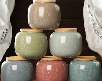 May include: Six ceramic storage jars with wooden lids. The jars are in various colors, including gray, green, brown, and blue. The jars are arranged in a pyramid shape.