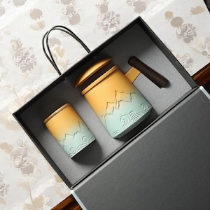 以下が含まれることがあります： 黒いギフトボックスに入った、蓋付きのセラミック製ティーカップ2個セット。カップは、黄色と青の色合いで山と波のデザインが施されています。