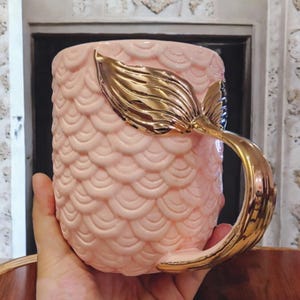 May include: A pink ceramic mug with a gold mermaid tail handle. The mug is decorated with a mermaid scale pattern.