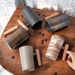 May include: Four ceramic mugs with wooden handles. The mugs are in different colors and patterns, including black, brown, beige, and white. The mugs are arranged on a wooden surface.