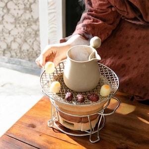 May include: A white ceramic teapot with a wooden handle sits on a small metal grill over a clay stove. The stove is on a wooden table. There are small white and brown treats on the grill.