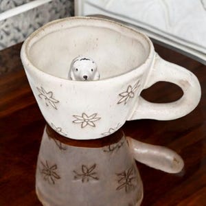 May include: White ceramic mug with a small white ceramic animal figurine inside. The mug has a floral pattern and a handle.