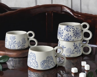 May include: Set of four white ceramic mugs with blue floral designs. Each mug has a unique design and a double handle. The mugs are arranged on a wooden surface with sugar cubes.
