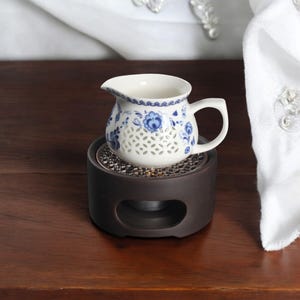 Puede incluir: Una tetera de cerámica blanca con un diseño floral azul y blanco se asienta sobre un soporte de calentamiento de cerámica negro. La tetera tiene un pequeño pico y una manija.