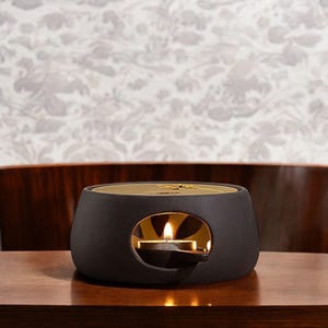 May include: Black ceramic tea warmer with a gold top and a small candle inside. The warmer is on a wooden surface.