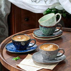 Puede incluir: Tres tazas de café de cerámica con arte latte sobre una mesa de madera. Las tazas son de diferentes tonos de azul, verde y gris. Cada taza tiene un platillo a juego.