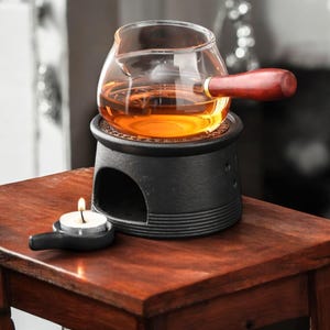 Puede incluir: Una tetera de vidrio con asa de madera se asienta sobre un calentador de hierro fundido negro con una vela de té debajo. La tetera está llena de un líquido dorado.