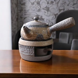 Puede incluir: Una tetera de cerámica marrón y gris con asa y pico, sentada sobre un calentador de cerámica a juego con una taza integrada.