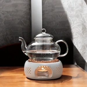 Puede incluir: Una tetera de vidrio transparente con un infusor incorporado se encuentra sobre un calentador de té de cerámica blanca con un portavelas. El calentador tiene un patrón decorativo y una pequeña vela está encendida en su interior.