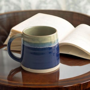 Può includere: Una tazza in ceramica blu e verde con un bordo bianco. La tazza è appoggiata su un tavolo di legno accanto a un libro.