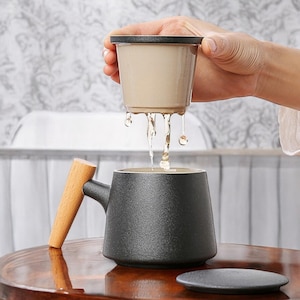 May include: A black ceramic tea mug with a wooden handle and a matching saucer. The mug has a textured surface and is being filled with tea from a beige ceramic strainer.