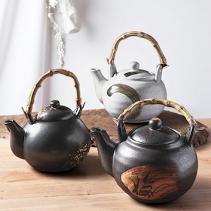 Puede incluir: Tres teteras de cerámica con asas de bambú. Las teteras son de diferentes colores y tienen diferentes diseños. Una es blanca con un diseño de remolino gris, otra es negra con un diseño moteado y otra es negra con un diseño de hoja marrón.