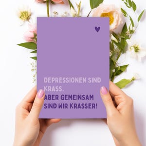 Könnte beinhalten: Eine lila Karte mit einem lila Herzsymbol wird von Händen mit rosa Nagellack gehalten, umgeben von Blumen. Auf der Karte steht der Text: "DEPRESSIONEN SIND KRASS. ABER GEMEINSAM SIND WIR KRASSER!"