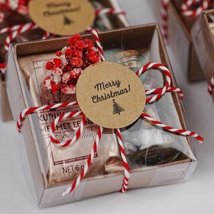 May include: Four small clear boxes with brown lids, each filled with a small gift and decorated with red and white twine, red dried flowers, and a round brown paper tag with the text "Merry Christmas!"