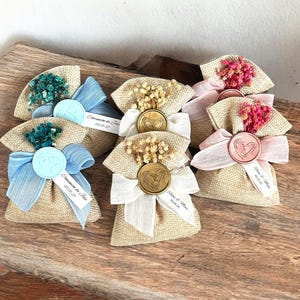 May include: Assortment of small burlap bags with different colored ribbons and dried flowers. Each bag has a wax seal with a heart design and a personalized tag with a name and date.