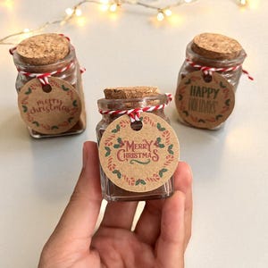 Op de afbeelding: Kleine glazen potjes met kurken deksels, gevuld met een donker poeder. Elk potje heeft een rond label met vakantiegroeten: "Merry Christmas" of "Happy Holidays". De labels zijn vastgebonden met rood en wit touw.