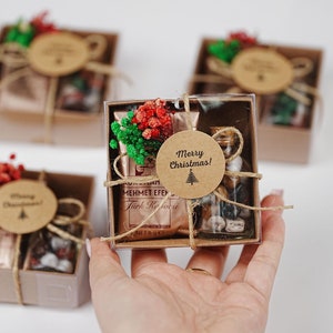 May include: Four small brown gift boxes tied with twine and decorated with a sprig of greenery and a small red flower. Each box has a round brown tag with the text "Merry Christmas!" and a small Christmas tree graphic. The boxes contain small gifts, such as a bottle of spices and a bag of tea.