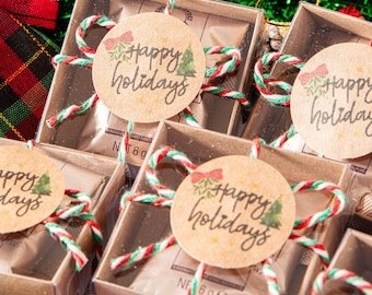 May include: Several small gift boxes with clear lids are arranged, each adorned with a round tag that reads "happy holidays." The boxes are tied with red and green twine. Festive decorations, including a plaid bow and candy canes, are in the background.