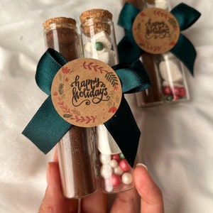 Pu&ograve; includere: Due tubi di vetro riempiti con miscela per cioccolata calda, marshmallow e codette. I tubi sono legati insieme con un nastro verde e un'etichetta di carta marrone che dice "Buone Feste".