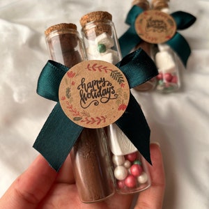 Pu&ograve; includere: Due provette di vetro riempite con miscela per cioccolata calda, marshmallow e codette. Le provette sono legate insieme da un nastro verde e un'etichetta circolare di carta marrone che dice "Buone Feste".