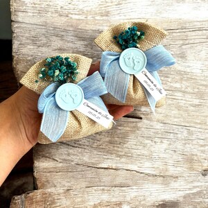 May include: Two small burlap bags with blue ribbon and a blue wax seal with a footprint design. Each bag has a small tag with the text "Comunione de Jose" and the date "08.03.25".