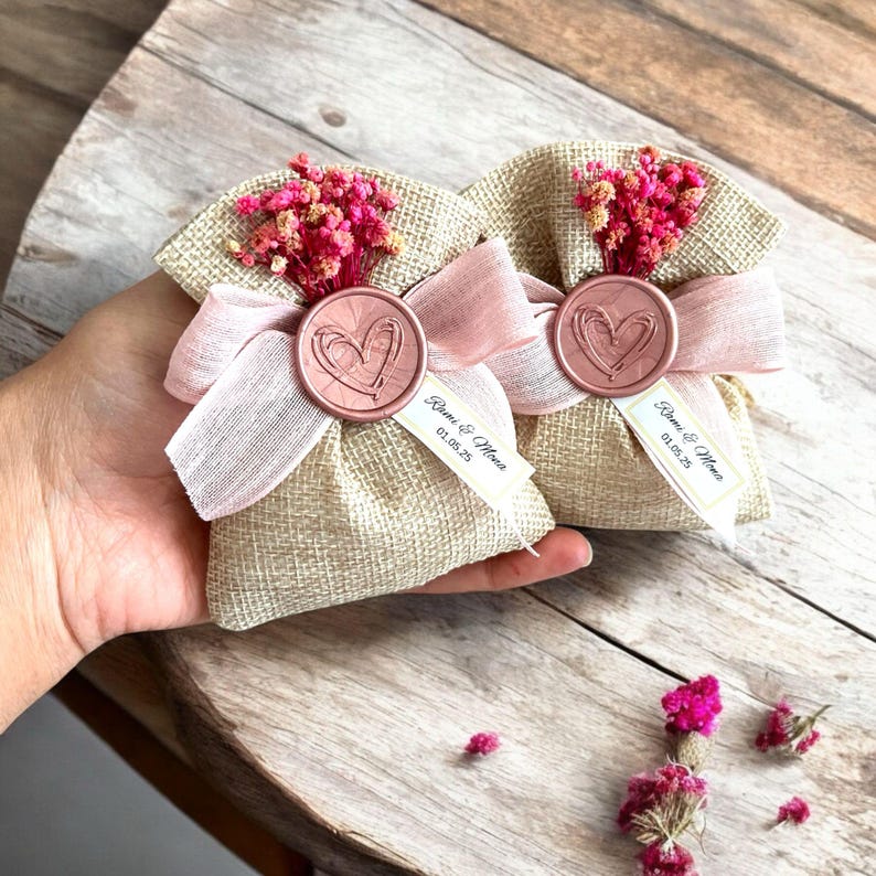 May include: Two small burlap bags with pink ribbon bows and a rose gold wax seal with a heart design. Each bag has a small white tag with the text "Rini & Mona 01.05.23".