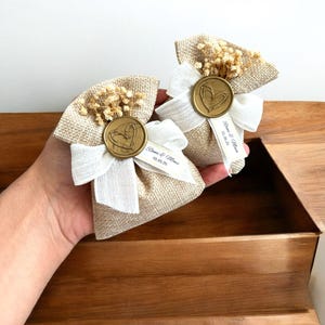 May include: Two small burlap bags with white ribbon bows and dried flowers. Each bag has a gold wax seal with a heart design and a personalized label with the text "Roma & Mona 01.05.25".