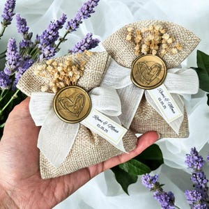 May include: Two small burlap bags with white ribbon bows and a gold heart wax seal. Each bag has a small white tag with the text "Rami & Mona 01.05.25".