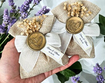 Puede incluir: Dos pequeñas bolsas de arpillera con lazos de cinta blanca y un sello de cera en forma de corazón dorado. Cada bolsa tiene una pequeña etiqueta blanca con el texto "Rami & Mona 01.05.25".