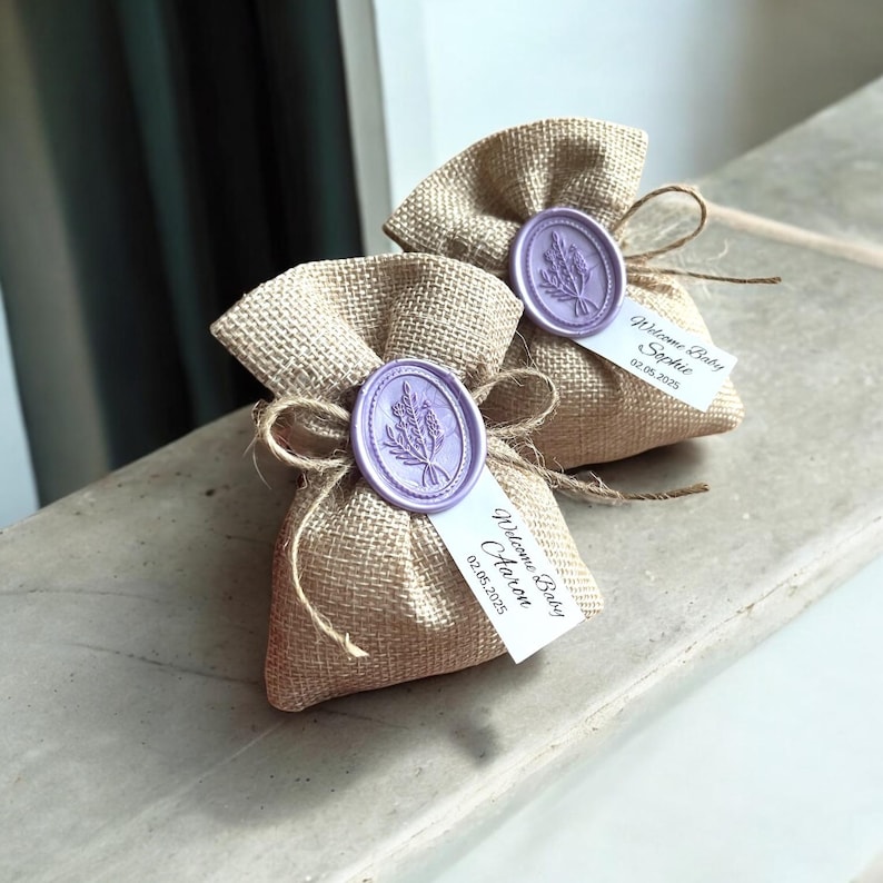 May include: Two small burlap bags with lavender-colored wax seals and personalized tags. The tags read "Welcome Baby Sophie 03.05.2025" and "Welcome Baby Aaron 02.08.2025".