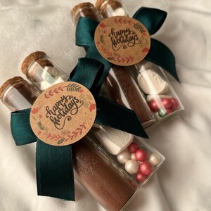 Pu&ograve; includere: Tre tubi di vetro legati con un nastro verde e un'etichetta marrone che dice "Buone Feste". Ogni tubo contiene miscela di cioccolata calda, marshmallow e codette rosse e bianche.