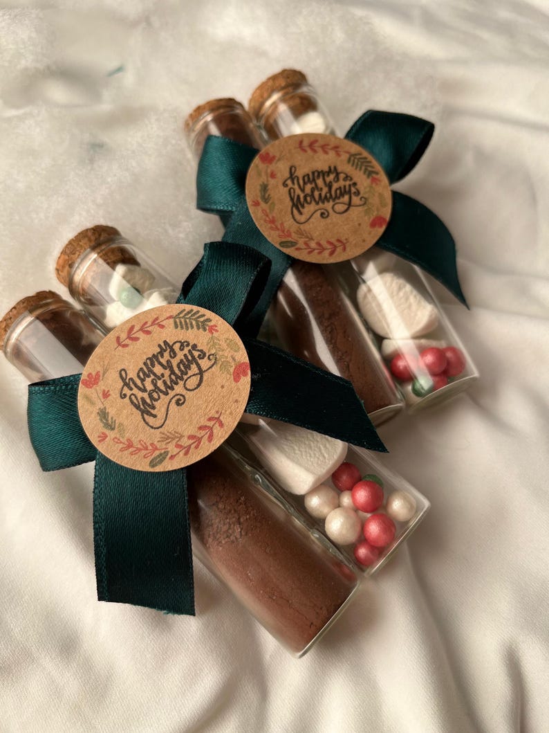 Pu&ograve; includere: Tre tubi di vetro legati con un nastro verde e un'etichetta marrone che dice "Buone Feste". Ogni tubo contiene miscela di cioccolata calda, marshmallow e codette rosse e bianche.