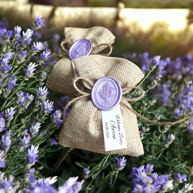 May include: Two small burlap bags with lavender-colored wax seals and a tag that reads "Welcome Baby,  Otaron, 02.05.2025". The bags are surrounded by lavender flowers.