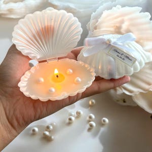 Puede incluir: Portavelas blanco en forma de concha con una pequeña vela blanca en su interior. El portavelas está abierto y la vela está encendida. Hay pequeñas perlas blancas esparcidas alrededor del portavelas.