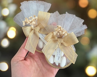 May include: Two small, round, gold-rimmed containers filled with white candy. Each container is wrapped in a sheer white tulle and a gold ribbon with a bow. The tulle is tied with a small bouquet of white dried flowers. Each container has a small white tag with black text that reads "Rina & Alvino 01.02.23".
