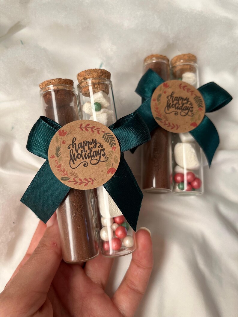 Pu&ograve; includere: Due provette di vetro riempite con miscela per cioccolata calda, marshmallow e codette. Le provette sono legate insieme da un nastro verde e un'etichetta di carta marrone che dice "Buone Feste".