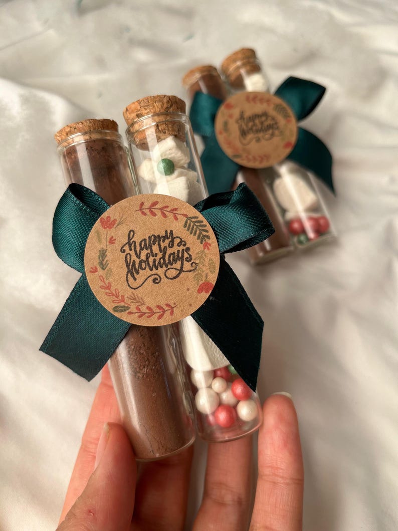 Pu&ograve; includere: Due provette di vetro riempite con miscela per cioccolata calda, marshmallow e caramelle rosse e bianche. Le provette sono legate insieme da un nastro verde e un'etichetta circolare di carta marrone che dice "Buone Feste".