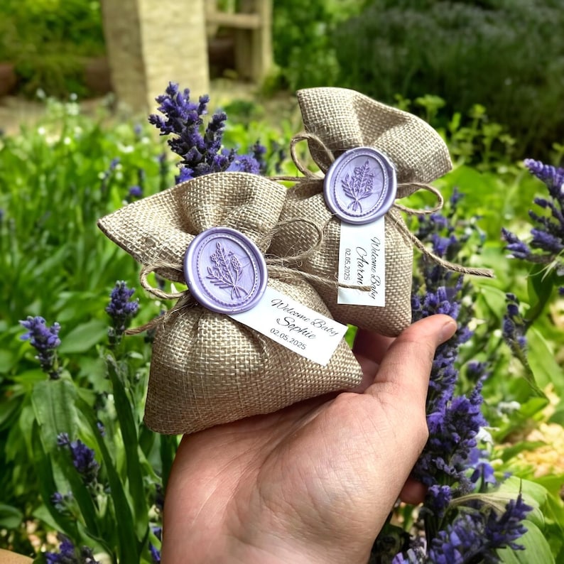 May include: Two small burlap bags with lavender-scented sachets. Each bag has a wax seal with a lavender sprig design and a personalized tag with the text "Welcome Baby Sophie 02.05.2023" and "Welcome Baby  Arianna 02.05.2023".