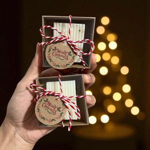 May include: Two small gift boxes each containing a bar of soap, tied with red and white twine. Each box has a round tag that says "Merry Christmas" in a festive font. The soap is a light color, and the boxes are brown.
