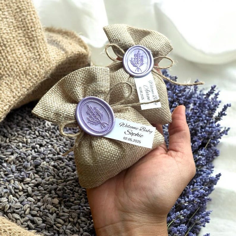 May include: Two small burlap bags with purple wax seals and personalized tags. The tags say "Welcome Baby Olivia 02.06.2025" and "Welcome Baby Sophie 02.05.2025". The bags are filled with lavender.