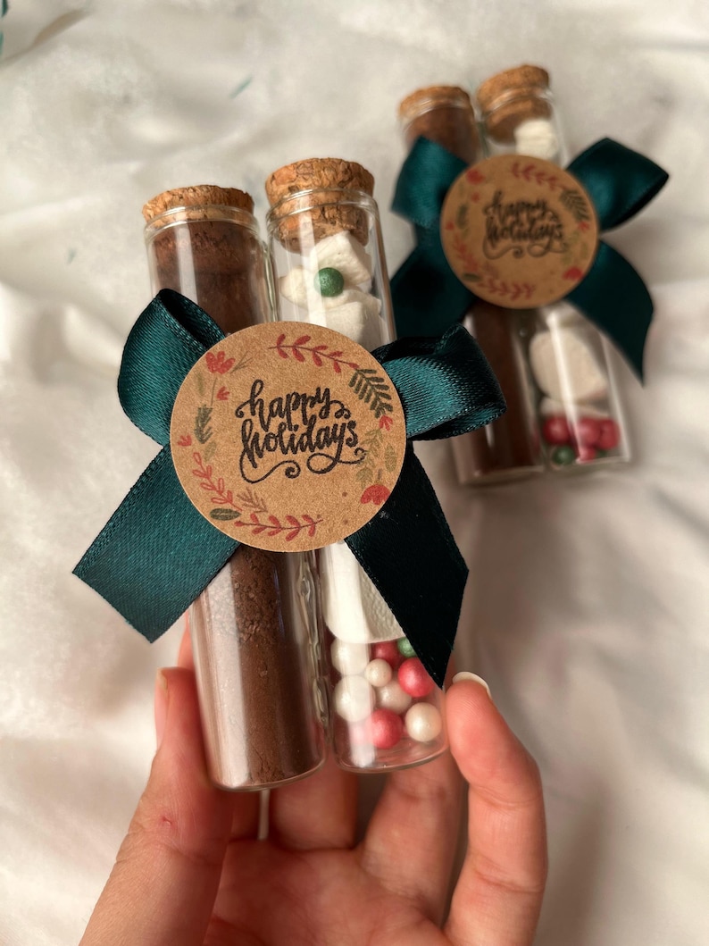 Pu&ograve; includere: Due tubi di vetro riempiti con miscela per cioccolata calda, marshmallow e codette. I tubi sono legati insieme con un nastro verde e un'etichetta di carta marrone che dice "Buone Feste".