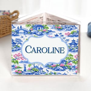 May include: A clear acrylic triangular table sign featuring a blue and white chinoiserie-style landscape pattern with pink floral accents. The center displays a white scalloped label with the name CAROLINE printed in dark blue capital letters.