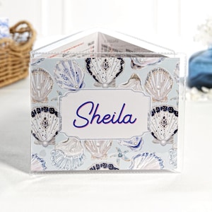 May include: A triangular clear acrylic table sign featuring a light blue background with a repeating pattern of white and navy blue seashells and pearls. A central white rectangular label displays the name Sheila in blue script lettering.