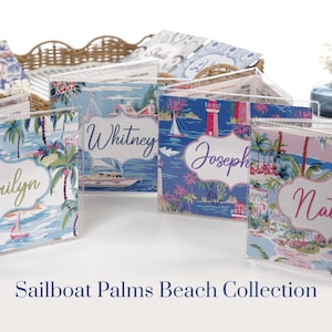 Bright blue and hot pink beach coastal Mahjong card cover collection with personalized names, displayed in clear sleeves on a white tabletop. Four colorful designs feature sailboats, palm trees, tropical waterfront scenes, beach houses, and a lighthouse in aqua, navy, blush pink, hot pink, green, and white for large 2025/2026 NMJL Mahjong cards.