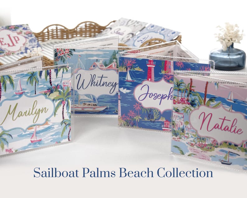 Bright blue and hot pink beach coastal Mahjong card cover collection with personalized names, displayed in clear sleeves on a white tabletop. Four colorful designs feature sailboats, palm trees, tropical waterfront scenes, beach houses, and a lighthouse in aqua, navy, blush pink, hot pink, green, and white for large 2025/2026 NMJL Mahjong cards.