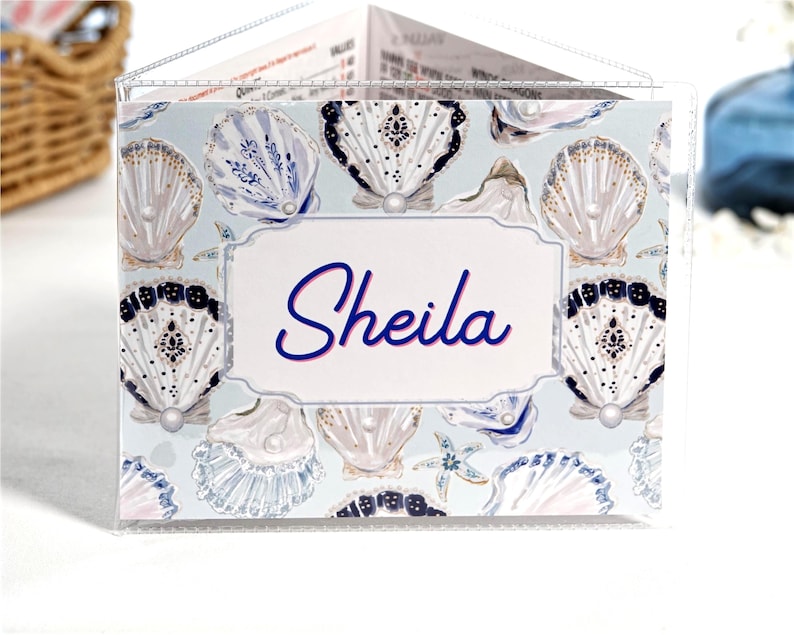 Pearl Shells personalized mahjong card cover for Large NMJL cards, shown in a clear sleeve with the name Sheila, featuring ocean beach coastal seashell artwork in soft blue, cream, navy, gold, pearl, and blush tones as a custom mahjong card holder and Mah Jongg accessory.